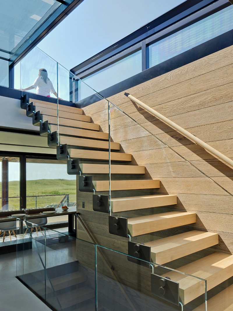 Moderne Treppe mit Holzstufen und Glasgeländer in einem hellen, minimalistischen Raum. Im Hintergrund ist eine Person zu sehen, die eine Glaswand hochgeht. Große Fenster lassen viel Tageslicht herein.