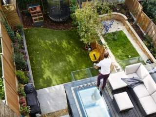 Vue aérienne d'un jardin aménagé avec du gazon et des plantes. Un homme se tient sur une terrasse, près d'un canapé moderne. On aperçoit un trampoline et un coin de jardin avec un petit banc et des plantations.