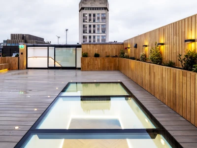 Une terrasse moderne avec un sol en bois, une piscine transparente et un bâtiment en arrière-plan sous un ciel nuageux. Des lampes éclairent la scène, créant une ambiance chaleureuse.