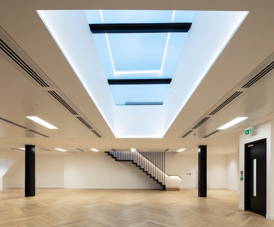Intérieur moderne avec un plafond lumineux et un puits de lumière. Un escalier en bois blanc mène à un niveau supérieur. Le sol est en parquet clair et les murs sont peints en blanc.