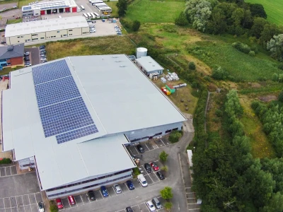 Vue aérienne d’un bâtiment industriel avec des panneaux solaires sur le toit, entouré de verdure et d’un terrain de stationnement. Des installations supplémentaires sont visibles à proximité, ainsi que des champs.
