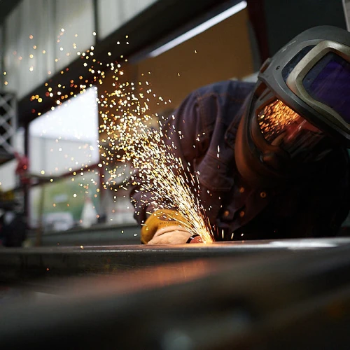 Un soudeur travaille sur un métal, produisant des étincelles brillantes. Il porte un casque de protection et des lunettes de sécurité, concentré sur sa tâche dans un atelier.