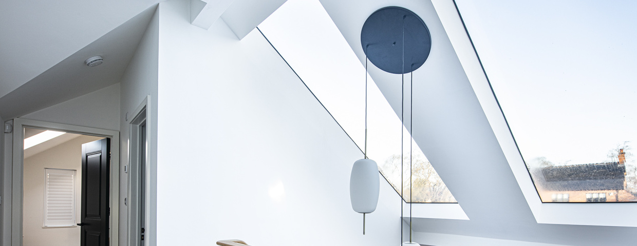 Vue d’un espace intérieur lumineux avec un grand plafond incliné. Des fenêtres situées dans le plafond laissent entrer la lumière naturelle. Un garde-corps en bois sépare l’escalier, et un luminaire suspendu apporte une touche moderne.