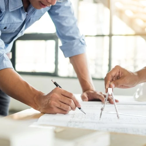 Deux personnes travaillent sur des plans architecturaux. L'une utilise un compas pour tracer des lignes, tandis que l'autre prend des notes avec un stylo. Un casque de sécurité est visible sur la table. La lumière naturelle entre par les fenêtres en arrière-plan.