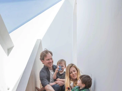 Une famille assise sur des escaliers en bois clairs. Les parents sourient, tenant chacun un enfant. L’un des enfants boit dans une bouteille, tandis que l’autre regarde. L’intérieur est moderne avec des murs blancs et un éclairage naturel.