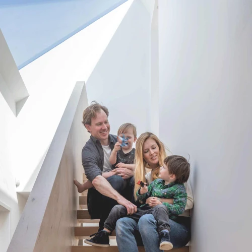 Une famille assise sur des escaliers en bois clairs. Les parents sourient, tenant chacun un enfant. L’un des enfants boit dans une bouteille, tandis que l’autre regarde. L’intérieur est moderne avec des murs blancs et un éclairage naturel.