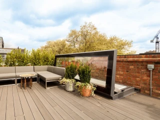 Une terrasse moderne avec un canapé gris, une table ronde, et des pots de plantes. En arrière-plan, des arbres feuillus et un mur en briques. Le ciel est partiellement nuageux.