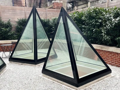 eux structures en forme de pyramide en verre, avec des bases noires, se trouvent dans une cour pavée de galets. En arrière-plan, on aperçoit des murs en briques et des fenêtres d’immeubles.
