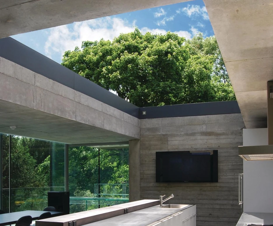 Cuisine moderne avec un grand plan de travail blanc, des armoires minimalistes et des fenêtres en verre laissant entrer la lumière naturelle. Un arbre verdoyant est visible à l’extérieur, créant une atmosphère harmonieuse avec l’espace.