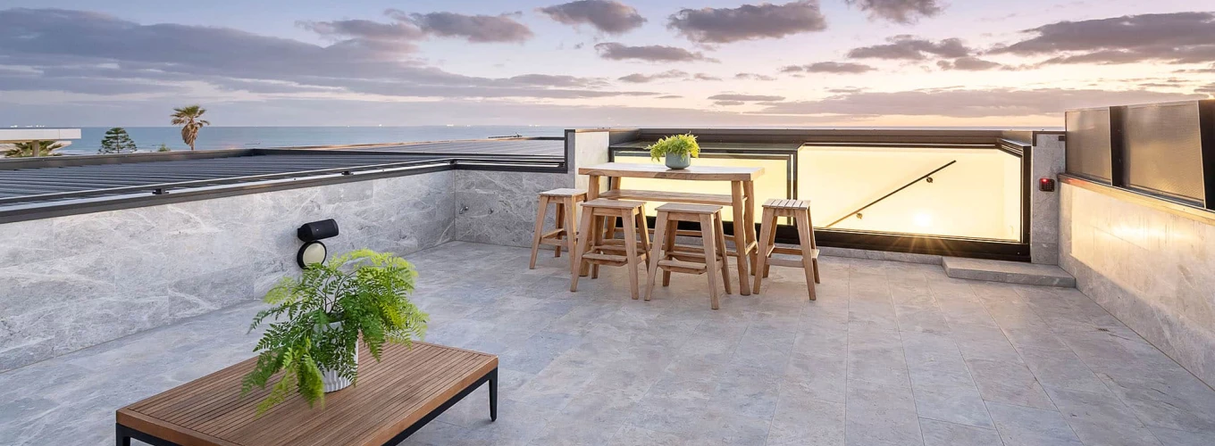 Terrasse spacieuse avec mobilier en bois, comprenant une table et des chaises, décorée de plantes. Le coucher de soleil se reflète sur l’horizon, créant une ambiance chaleureuse.