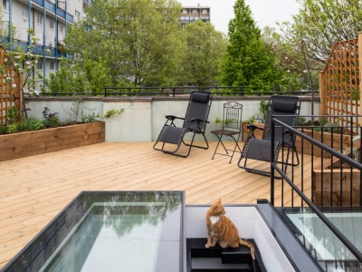 Terrasse en bois avec des chaises longues et une petite table. Un chat orange est assis au bord des escaliers menant à l'intérieur. Des arbres verdoyants entourent l'espace.