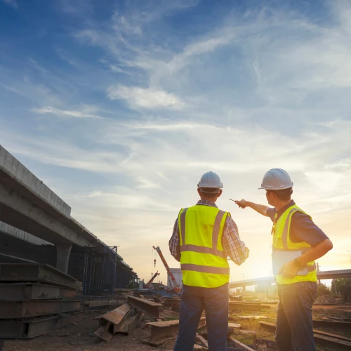 Deux travailleurs en chantier portent des casques et des gilets réfléchissants. Ils discutent et regardent un pont en construction au coucher du soleil, avec des structures en métal et des matériaux de construction visibles autour d'eux.