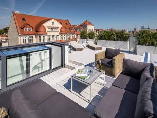 Terrasse moderne avec des canapés en rotin, une table basse et une vue sur un bâtiment de style architecture classique et la ville en arrière-plan. Le ciel est dégagé et ensoleillé.