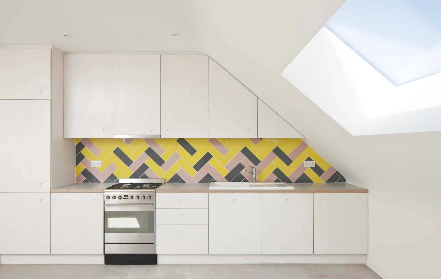 Cuisine moderne avec des armoires blanches, une hotte en inox, et un carrelage mural coloré en motifs zigzag jaune, rose et gris. Un puits de lumière illumine l'espace.