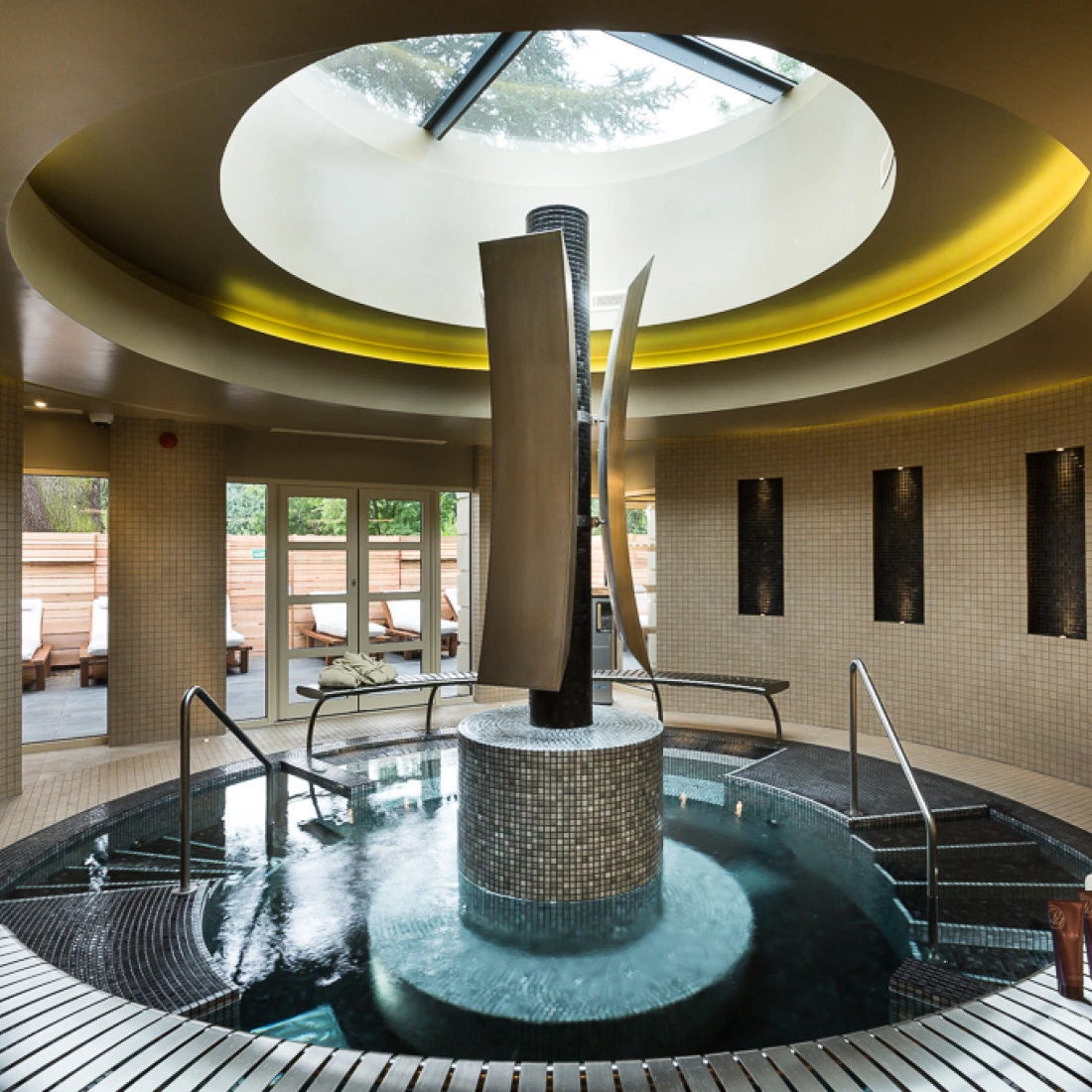 Un espace de spa moderne avec une grande piscine circulaire en mosaïque, entourée de murs en tuiles claires. Des colonnes métalliques élégantes s’élèvent au centre, tandis qu’un toit en verre laisse passer la lumière. Des chaises longues sont visibles à l’extérieur.