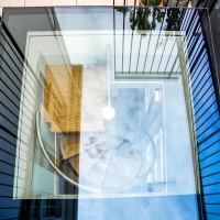 Vue plongeante sur un escalier en colimaçon en verre, entouré de structures noires, avec un reflet du ciel dans les surfaces vitrées. Un meuble en bois est visible à l'intérieur.
