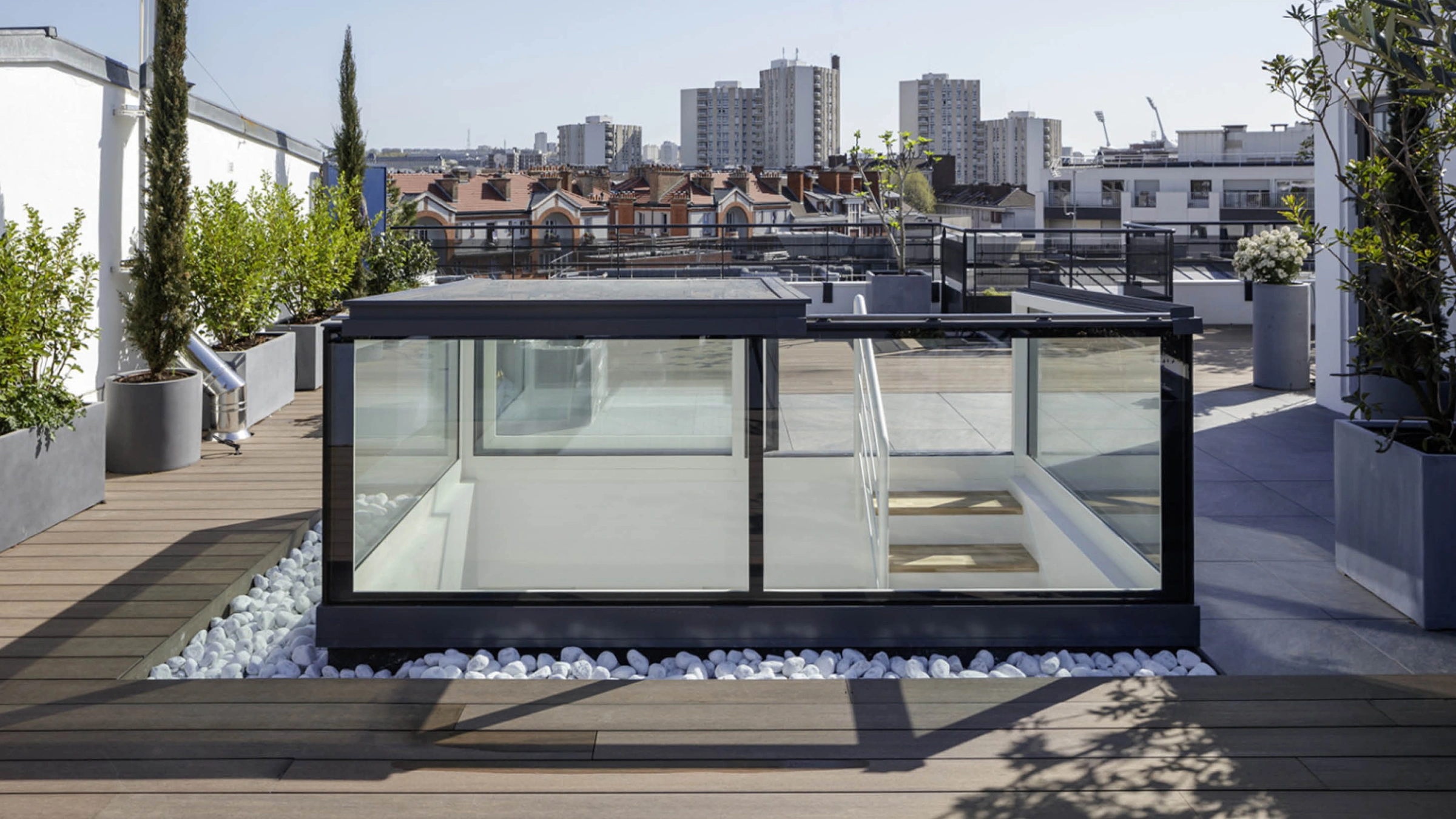 Un toit-terrasse moderne avec une structure en verre, entourée de plantes et de galets blancs. En arrière-plan, des bâtiments urbains sous un ciel bleu.