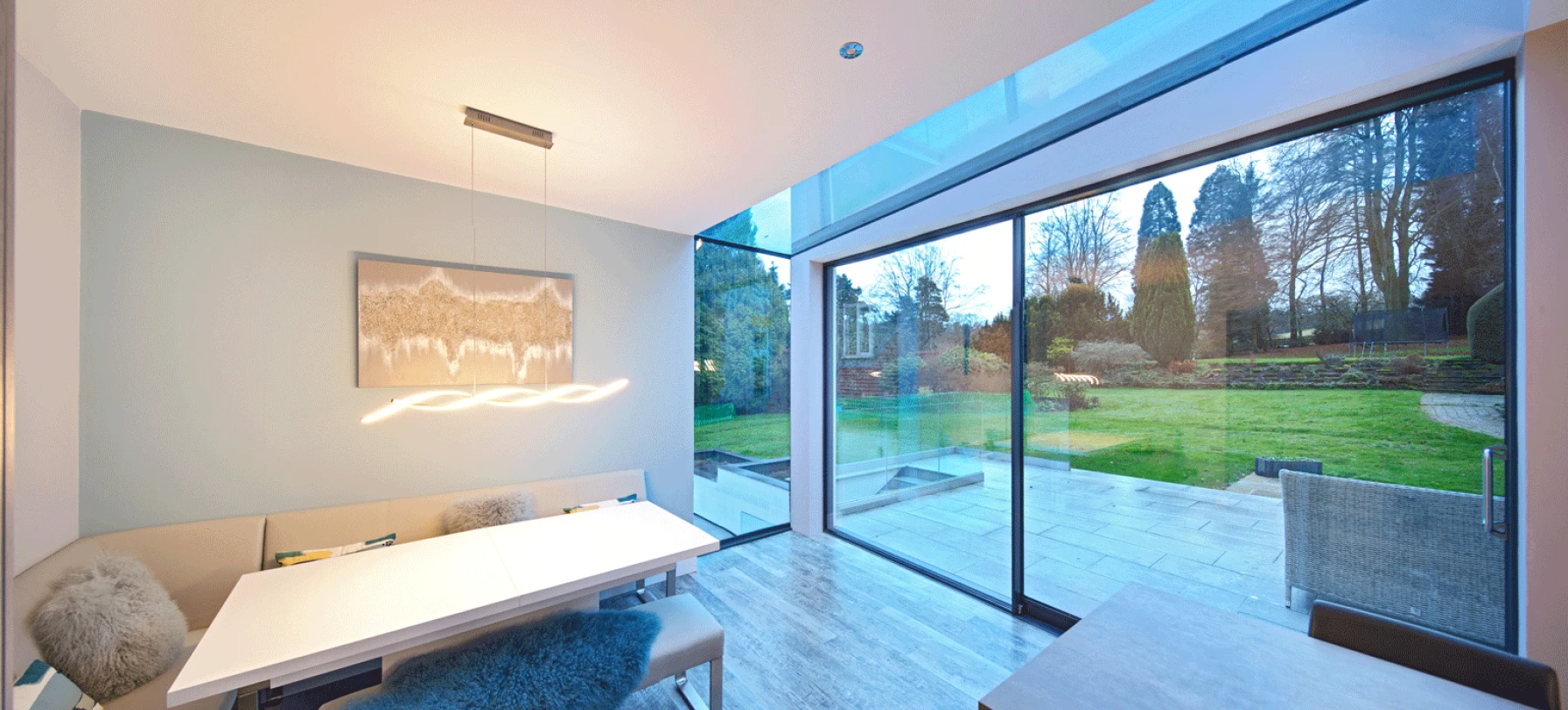 Un intérieur moderne d’une salle à manger avec une grande fenêtre donnant sur un jardin verdoyant. Une table blanche élégante est entourée de sièges confortables. Des lumières suspendues ajoutent une ambiance chaleureuse.
