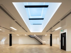 A modern, spacious room with light hardwood flooring and minimalist design. A large skylight in the ceiling allows natural light to enter, and a sleek staircase is visible in the background. Walls are painted white, enhancing the bright and airy atmosphere.