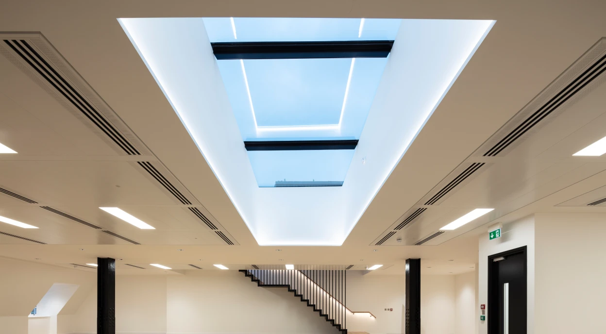Intérieur moderne avec un plafond lumineux et un puits de lumière. Un escalier en bois blanc mène à un niveau supérieur. Le sol est en parquet clair et les murs sont peints en blanc.