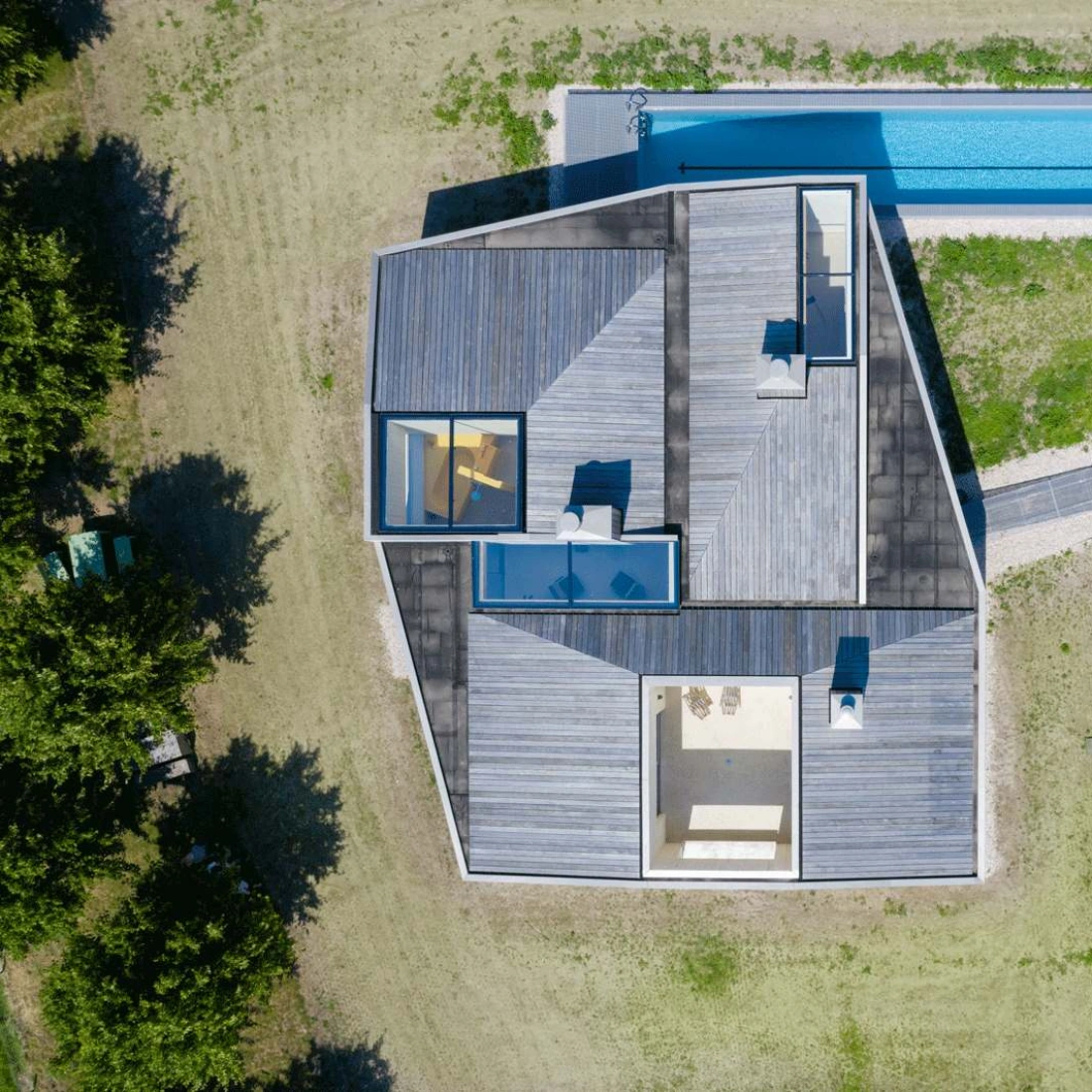 Vue aérienne d'une maison moderne avec un toit angulé, entourée d'un espace vert. Une longue piscine est visible à côté de la maison. Des chaises longues apparaissent sur la terrasse en bois.