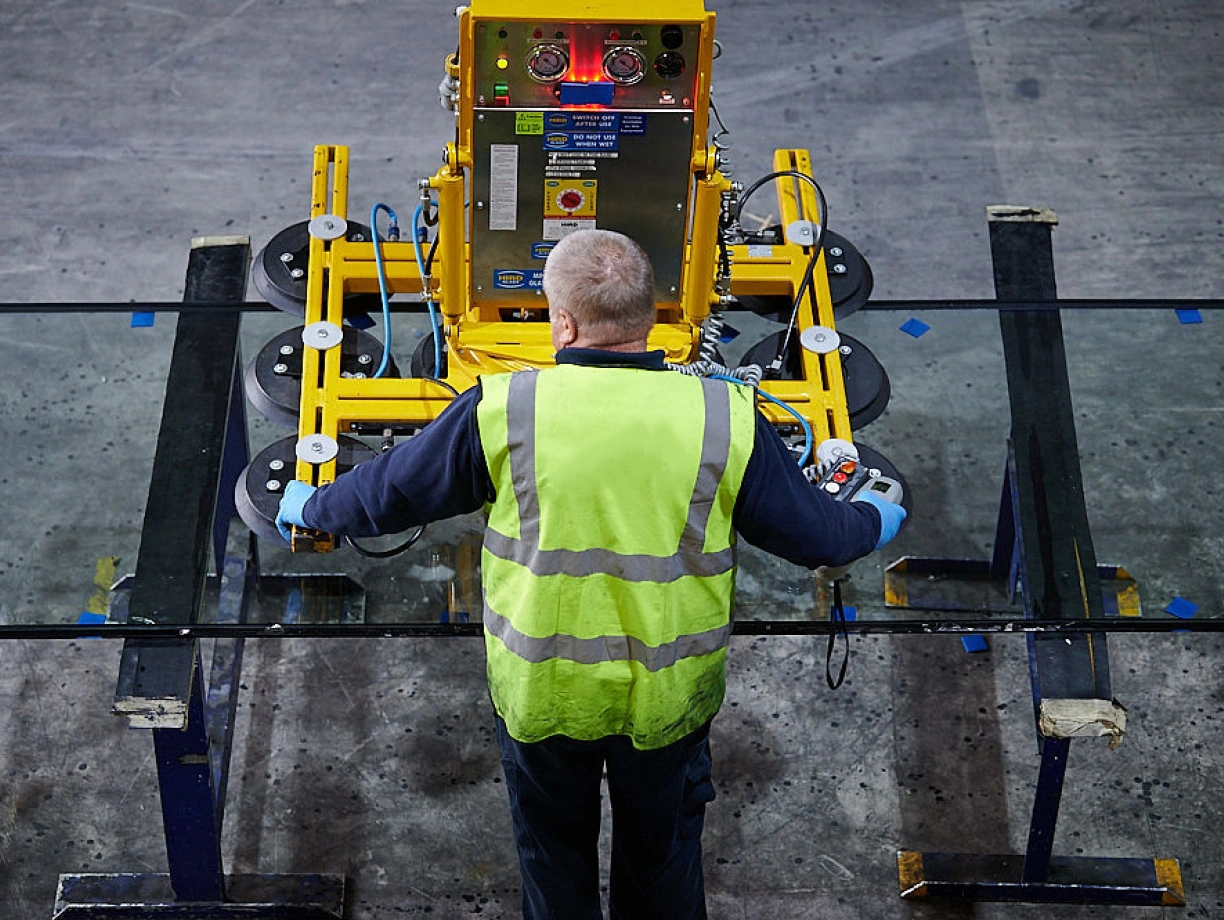 Un homme en gilet de sécurité utilise une machine jaune pour manipuler des panneaux de verre. L’atelier est vaste et le sol est en béton. Des lumières et des boutons sont visibles sur la machine.