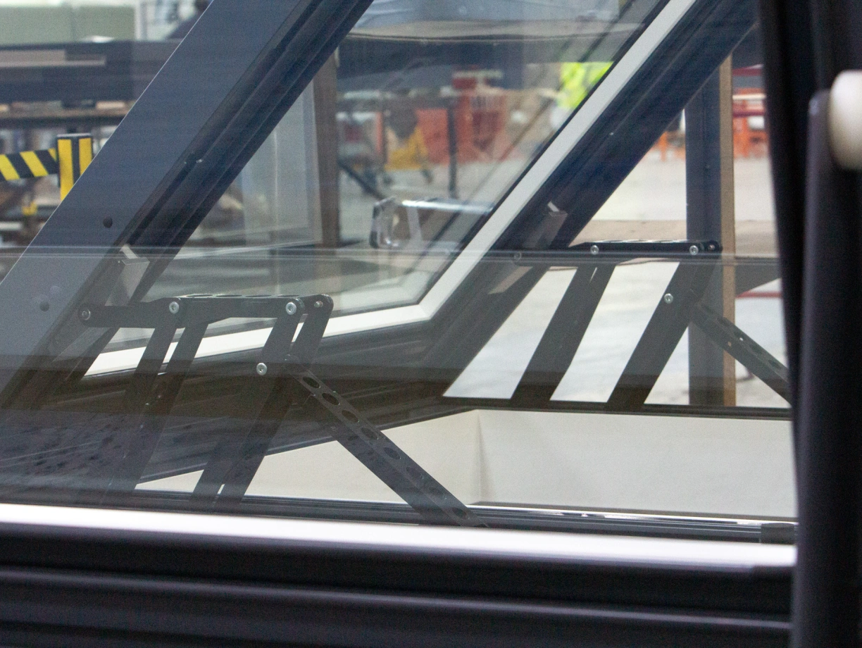 Vue rapprochée d’un système de support en métal à l’intérieur d’une structure en verre, avec des composants visibles et des travaux en arrière-plan.