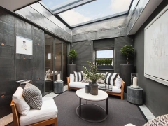 A modern sunroom featuring a sleek design with large skylights. Two sofas with white cushions and black decorative pillows are arranged around a round coffee table. The walls are a blend of dark materials, and there are potted plants and decorative elements enhancing the space. Natural light fills the room, creating a warm ambiance.
