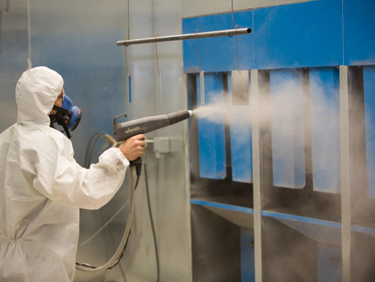 Un travailleur en combinaison de protection pulvérise de la peinture dans un espace de travail avec des murs bleus. De la brume de peinture s’échappe de l’outil.