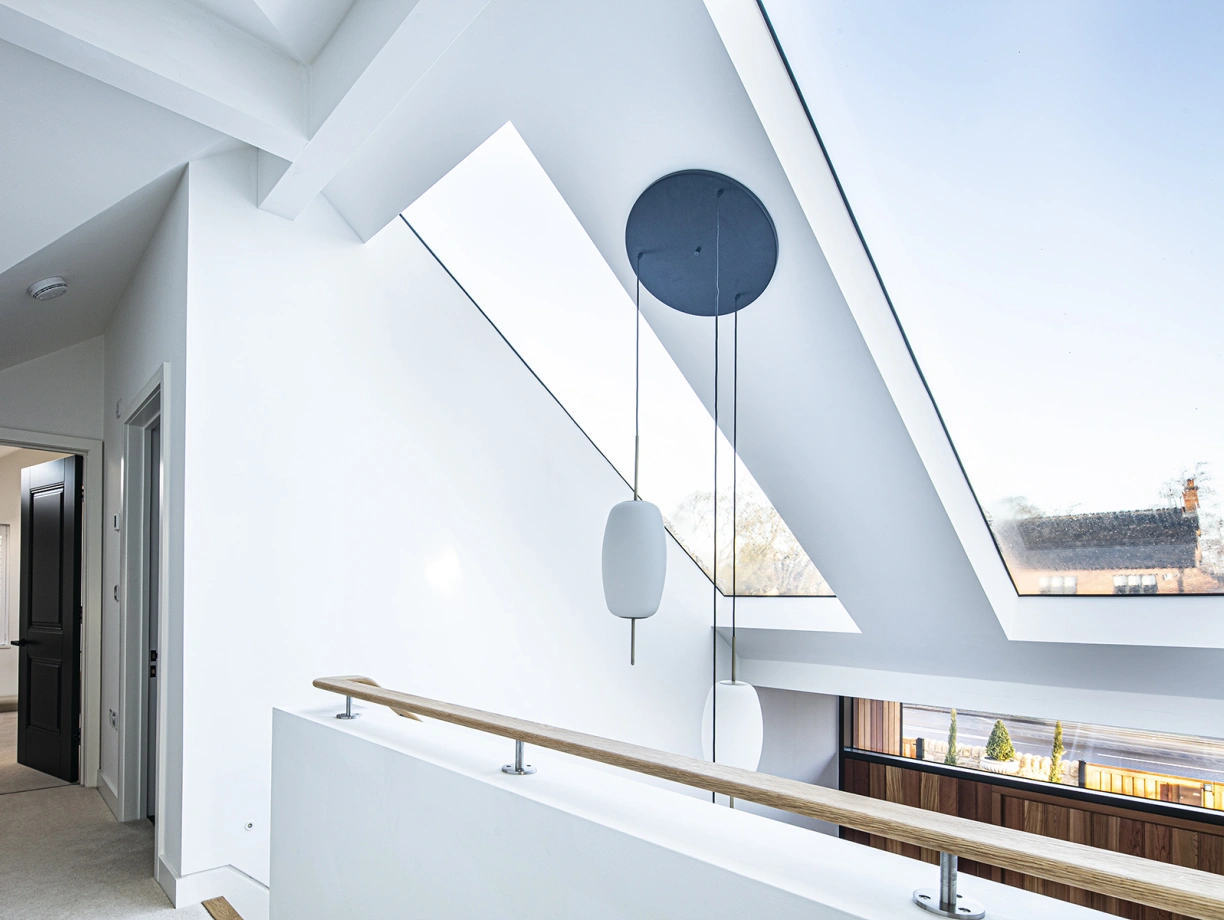 Vue d’un espace intérieur lumineux avec un grand plafond incliné. Des fenêtres situées dans le plafond laissent entrer la lumière naturelle. Un garde-corps en bois sépare l’escalier, et un luminaire suspendu apporte une touche moderne.