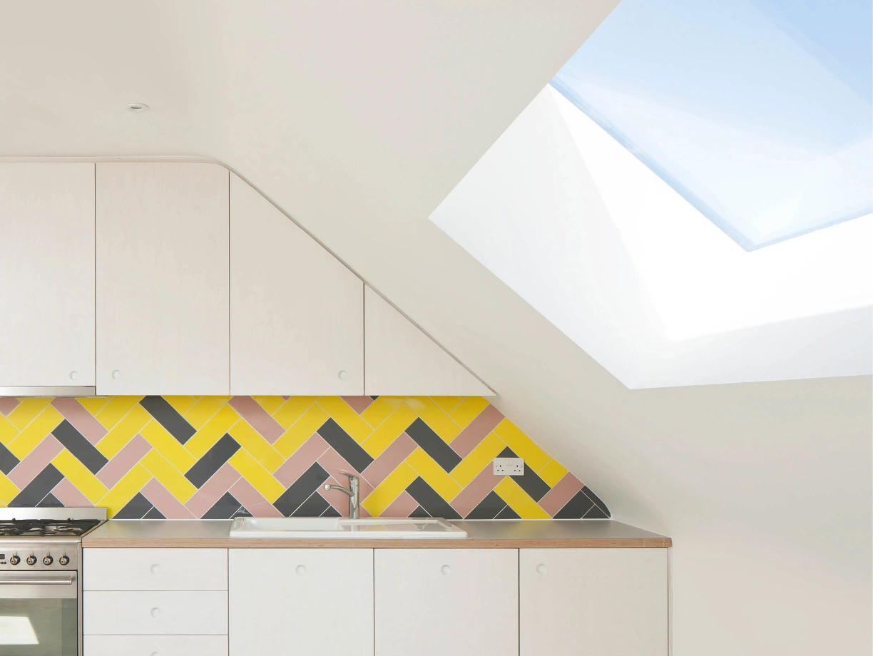 Cuisine moderne avec des carreaux en chevron jaune, rose et gris. Un four avec une hotte se trouve à gauche, tandis qu'un évier est installé sur un plan de travail en bois. Un puits de lumière au plafond illumine l'espace.