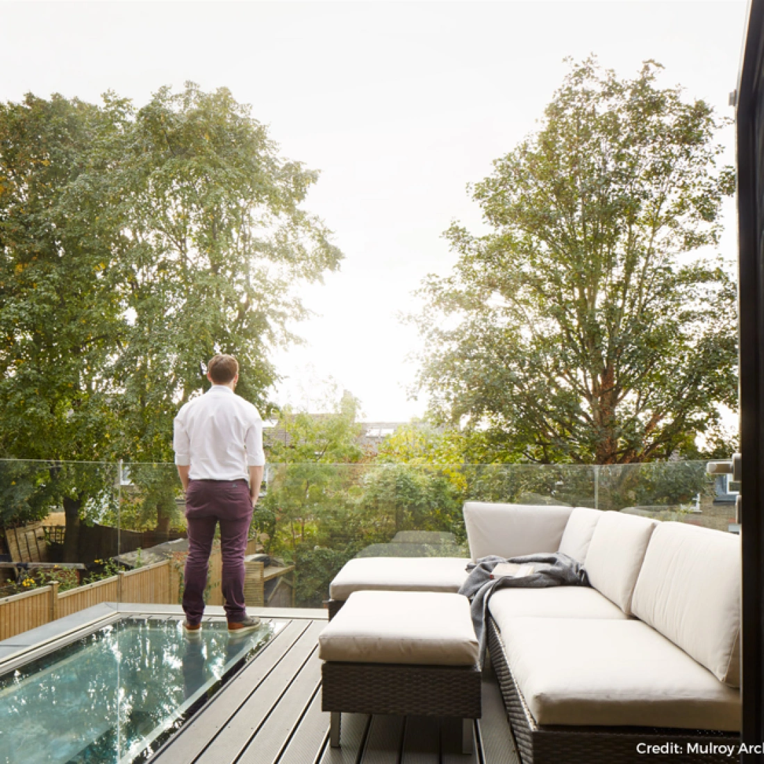 Un homme debout sur un balcon, regardant un paysage verdoyant avec des arbres. Il porte une chemise blanche et un pantalon violet. Un canapé moderne est visible à proximité.