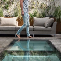 Un homme marche pieds nus sur un sol en verre à côté d'un salon d'extérieur moderne, entouré de plantes et de canapés en tissu.