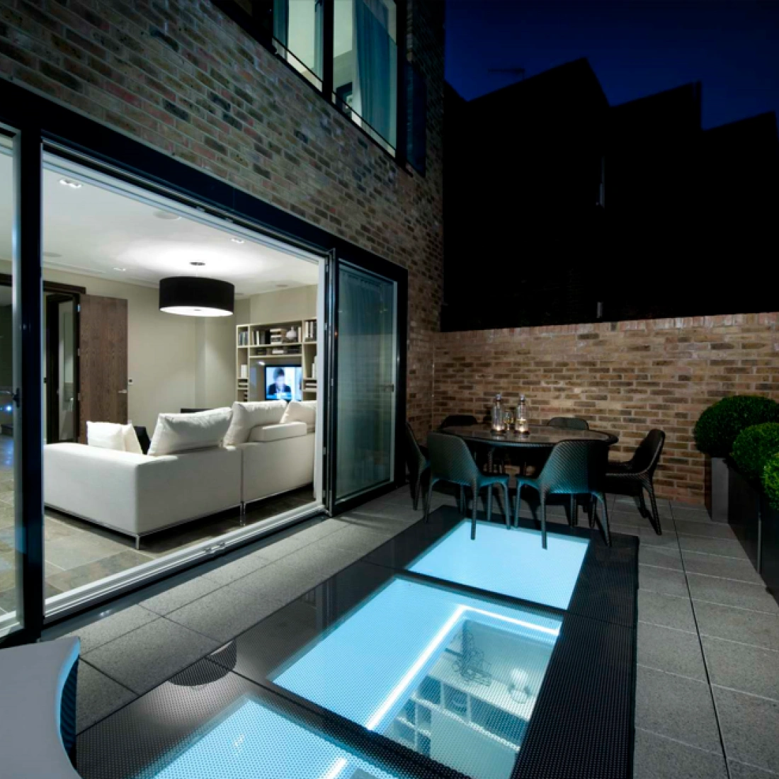 Terrasse moderne avec un sol en verre illuminé, des canapés blancs par paires à l’intérieur et une table à manger à l’extérieur. Le ciel nocturne crée une ambiance intime.