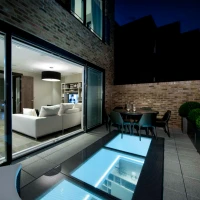 Terrasse moderne avec un sol en verre illuminé, des canapés blancs par paires à l’intérieur et une table à manger à l’extérieur. Le ciel nocturne crée une ambiance intime.