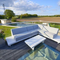 Un espace de détente extérieur avec un canapé gris en forme de L et une table basse blanche. En arrière-plan, un paysage de champs verdoyants sous un ciel nuageux.