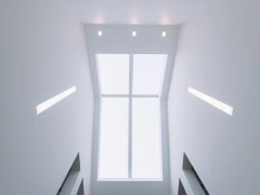 A view looking upward at a bright, minimalist stairwell. The ceiling is mostly white, featuring recessed lighting and a large rectangular window that lets in natural light.