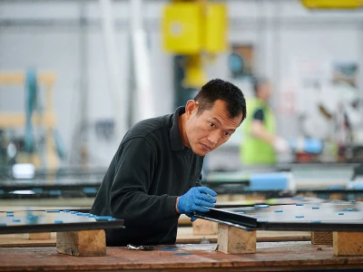 Un uomo lavora in una fabbrica, concentrato su un pannello di vetro che sta rifinendo. Indossa guanti blu e una maglietta scura. Sullo sfondo, si intravedono altre persone e attrezzature da lavoro.
