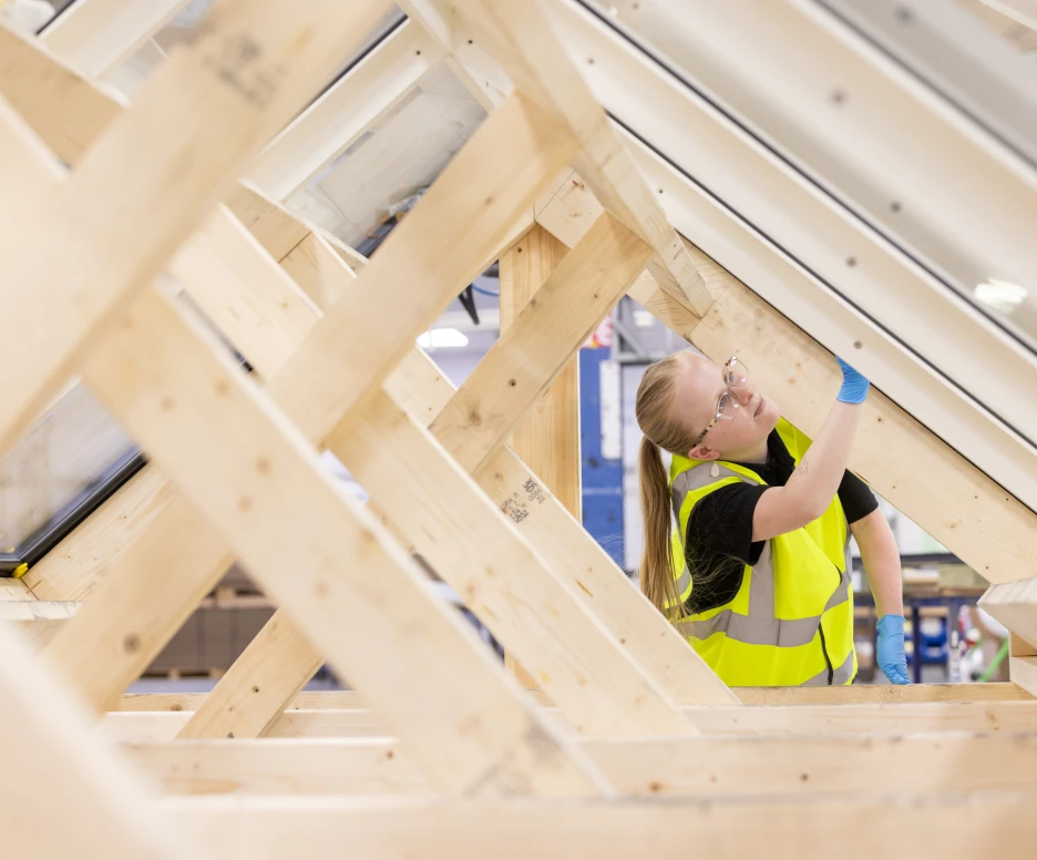 Un joven trabajando en una estructura de madera, ajustando un componente en un proyecto de construcciรณn. Lleva un chaleco reflectante y guantes. La luz entra por ventanas en el techo.