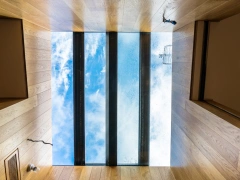 A view from below a glass ceiling, showcasing a clear blue sky with fluffy clouds and a hint of water droplets on the glass, framed by wooden walls.