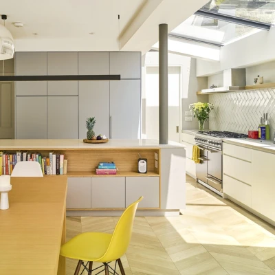 A modern kitchen featuring a light wooden dining table with two candles, a yellow chair, and a sleek island with storage. The kitchen has gray cabinetry, a white countertop, and stainless steel appliances, with a large window letting in natural light.