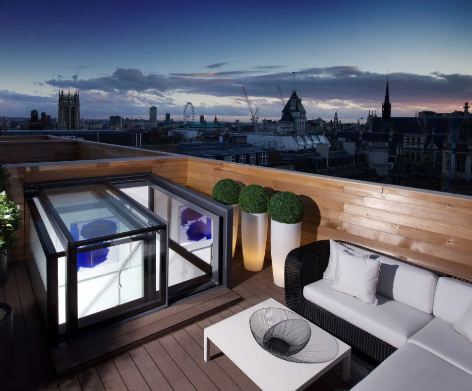 A stylish rooftop terrace features a modern seating area with a black wicker sofa and white cushions, alongside a sleek glass coffee table. A transparent hot tub is set within a black enclosure, with potted plants enhancing the decor. The backdrop showcases a breathtaking city skyline under a twilight sky.