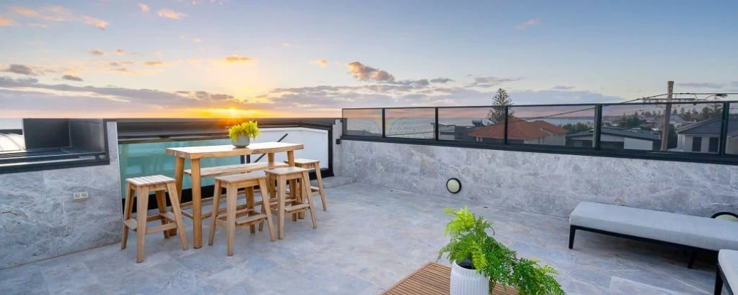 A rooftop terrace features a wooden dining table and chairs, a low wooden coffee table, and a potted plant. The scene is set against a vibrant sunset, illuminating the sky with shades of orange and pink. Surrounding buildings and a swimming pool are visible in the background.