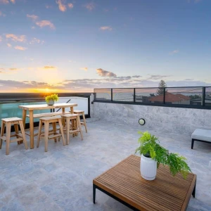 A rooftop terrace features a wooden dining table and chairs, a low wooden coffee table, and a potted plant. The scene is set against a vibrant sunset, illuminating the sky with shades of orange and pink. Surrounding buildings and a swimming pool are visible in the background.