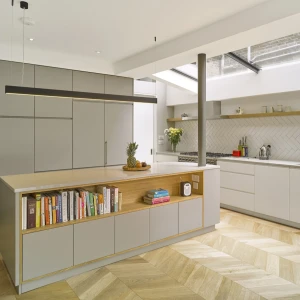 Modern kitchen featuring a large island with a wood-accented shelf displaying books, and sleek gray cabinetry. The space is illuminated by natural light from a skylight, with herringbone-patterned wood flooring and minimalist decor.