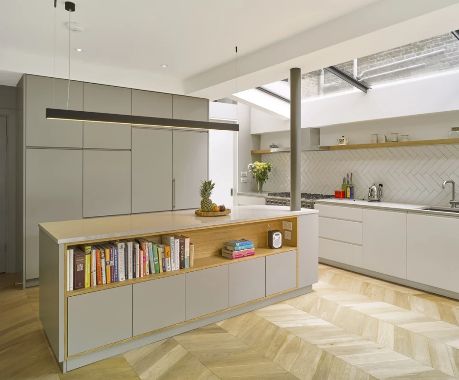 Modern kitchen featuring a large island with a wood-accented shelf displaying books, and sleek gray cabinetry. The space is illuminated by natural light from a skylight, with herringbone-patterned wood flooring and minimalist decor.