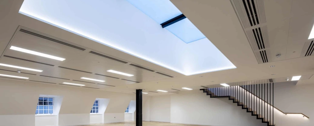 A spacious interior room featuring hardwood flooring and a minimalist design. Skylights illuminate the ceiling, which is adorned with modern lighting fixtures. A wooden staircase with a sleek railing is visible in the corner.