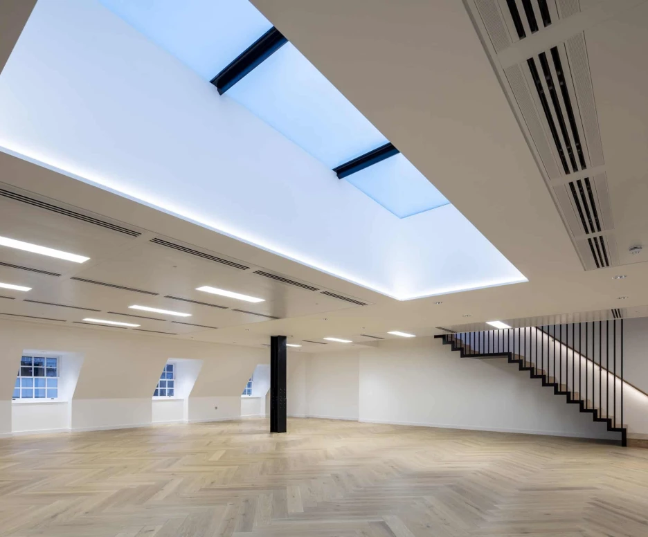 shows a stair way with a big empty room with a skylight in the middle of the room letting natural light shine in.