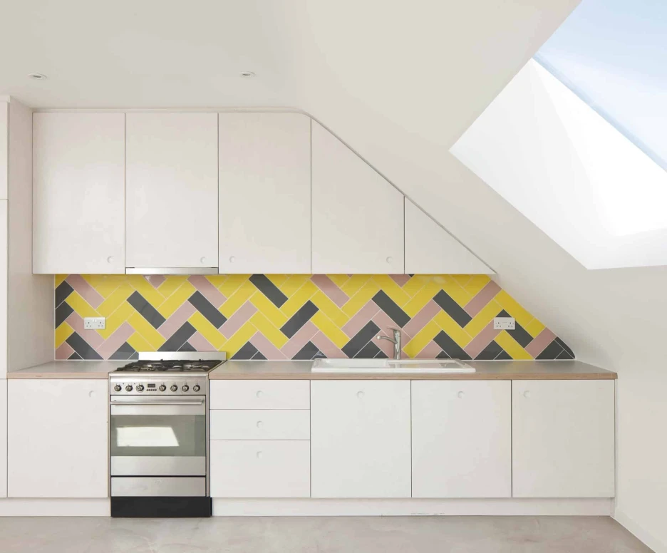 A white kitchen with a yellow, pink and purple back drop to the kitchen. To the right of the image there is a fixed Flushglaze skylight which is shining natural daylight into the kitchen.