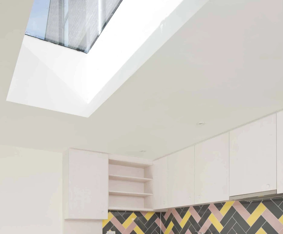 A modern kitchen featuring white cabinetry, a colorful herringbone tiled backsplash in pink, yellow, and gray, and a large skylight above that lets in natural light.